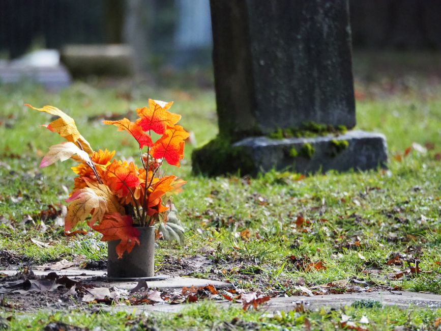 A floral arrangement of artificial autumn leaves in shades of orange, red, yellow, and light green, placed in a small, dark grey cylindrical vase on a grave's concrete base. The bouquet is positioned 