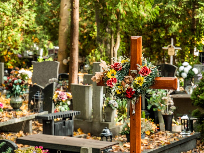 A wooden grave cross adorned with a floral arrangement consisting of red roses, yellow daisies, and pale pink flowers, surrounded by green foliage, positioned in a cemetery with numerous upright heads