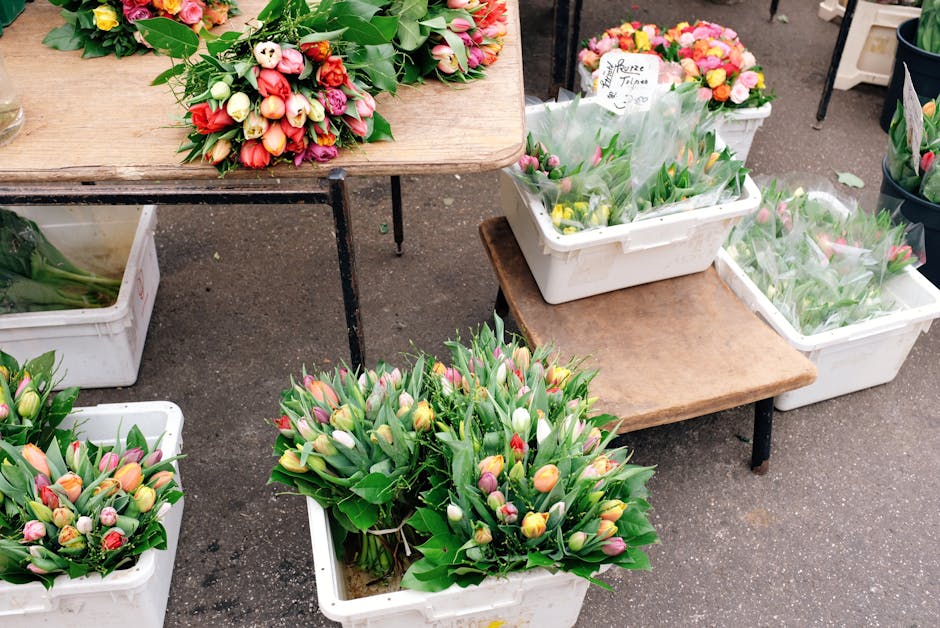 At a flower market stall, various bouquets of tulips and mixed spring flowers are arranged in white plastic crates and placed on wooden tables. The tulips exhibit pastel shades of pink, yellow, white,
