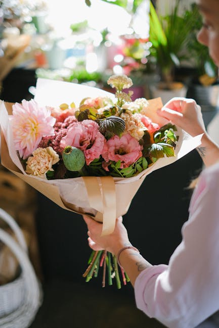 A person holding a large, vibrant floral bouquet composed of pink peonies, cream-colored carnations, and light purple dahlias, accented with green leaves and stems. The bouquet is wrapped in elegant c