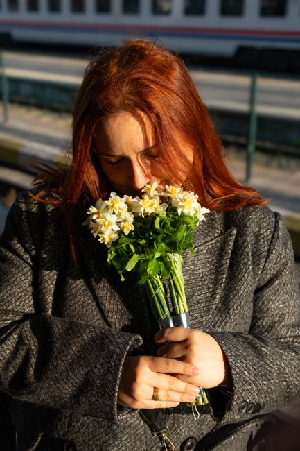 A woman with shoulder-length red hair, wearing a dark grey textured coat, is holding a bouquet of fresh daffodils with bright yellow petals and green stems wrapped in a simple transparent plastic slee