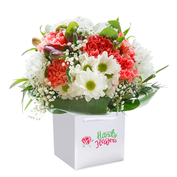This vibrant bouquet, showcased upright in a clear white gift bag, brings together the best of Northwood's floral artistry in a presentation designed to delight. The dominant coral and blush-toned carnations, with their opulent, frilled petals, are accented by fresh white daisy-like blooms whose yellow-green centers glow with optimism-an echo of Northwood's sunny green spaces and welcoming community spirit. Wisps of baby's breath ghost through the arrangement, lightening its form and adding gentle texture. Delicate closed buds tinged with soft purple nestle on the left, hinting at something new waiting to bloom. Lush, deep green foliage, tactile and lustrous, creates a supportive base that envelops the florals much as the woodlands surround Northwood's peaceful avenues. The modern white gift bag's neat ribbon handles signal ease and elegance, suited for sharing on memorable occasions or for doorstep delivery on Northwood's well-tended drives. A smart, locally branded emblem quietly affirms the bouquet's provenance. Each bloom is thoughtfully arranged to form a rounded cascade, projecting vitality, freshness, and heartfelt intent. Perfect for honouring a birthday, an anniversary, or simply a spontaneous act of kindness, this arrangement speaks of Northwood's love for thoughtful gifting and its rich floral tradition-offering a touch of celebration, artistry, and local pride.
