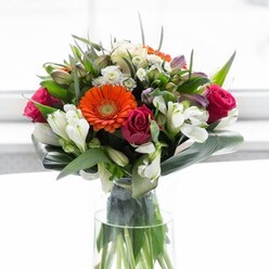 This artful, locally crafted bouquet sings with the colours and textures of a Northwood spring morning. Gentle peach and rich orange gerberas ring the arrangement with warmth and vitality, evoking the energy of Northwood's vibrant Marketplace or weekend strolls through Haste Hill Woods. Fuchsia and magenta roses nestle among creamy-white alstroemeria, each bloom contributing to the bouquet's multi-layered elegance and tactile appeal. Delicate hints of pale pink and petite white blossoms, possibly small spray roses or other fillers, provide subtle contrast and visual lift, enhancing the composition's sense of abundance and care. Thick, glossy aspidistra leaves curve around the stems at the base-a skilled florist's touch that both frames and supports the entire ensemble, underscoring the professionalism and thoughtfulness behind each arrangement made in Northwood. Sprigs of lush greenery fill the spaces between flowers, adding volume and dynamic texture in a natural, organic way. Set in a clear glass vase, the exposed green stems whisper of the arrangement's freshness and its quick journey from Flower Delivery Northwood's store to the recipient's door. The soft, diffused background draws you into the bouquet's vivid palette, inviting reflection, celebration, or a simple moment of joy-a floral centrepiece perfectly attuned to marking special occasions or welcoming someone to the neighbourhood.